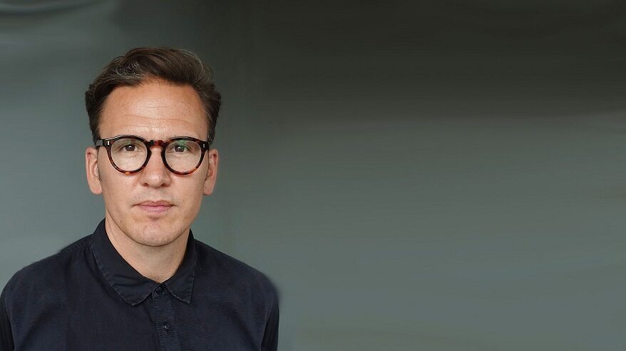 Close-up of Karl Warner, in dark shirt with glasses, against a dark background