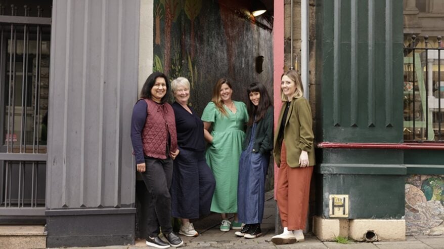 Four women stand against a building with two large grey pillars
