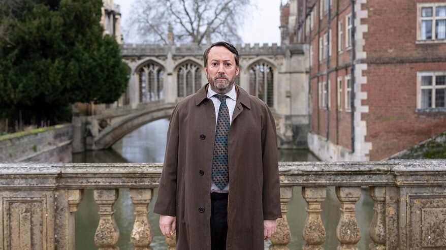 A man wearing a brown overcoat and smart clothes with tie stands on a stone bridge, with another old stone bridge and building behind