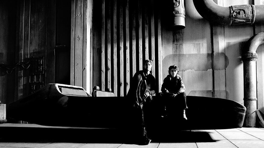 Black and white image of a man and a boy sitting together, against an industrial wall with giant pipes