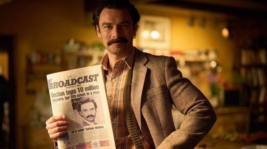 Aidan Turner in character in Disney+ 1980s-set drama Rivals, wearing fawn suit and holding a magazine