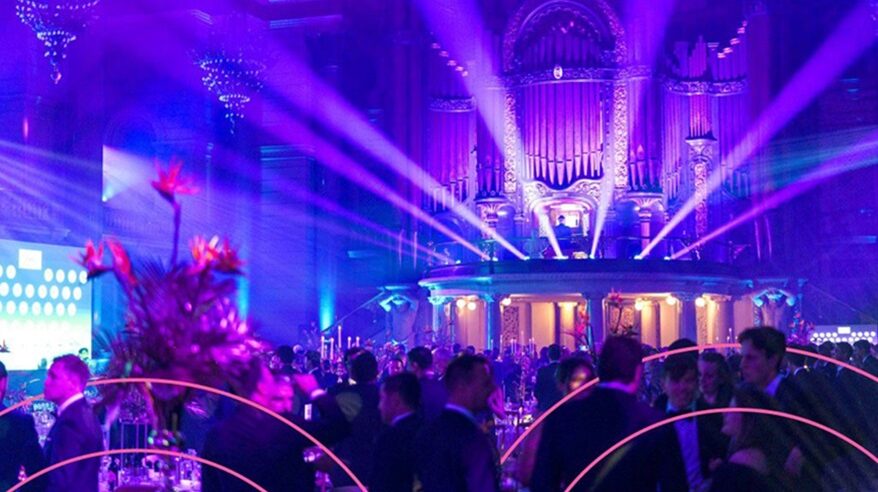 A glitzy, huge room with an organ, pink lighting and with people attending a function wearing tuxedos