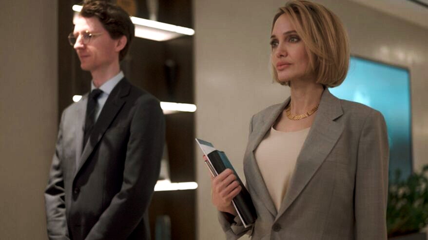 A woman in smart buff jacket holding a folder stands looking ahead. She has a blonde-ish bob and a man in a suit stands to her right in an office environment. From filming on movie Anxious People