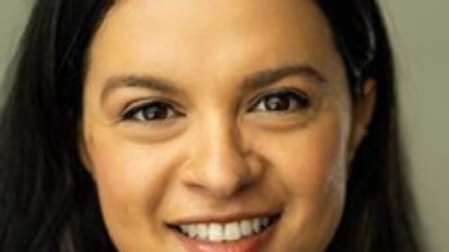 Headshot of a young woman with dark hair and centre parting looking straight to camera, smiling