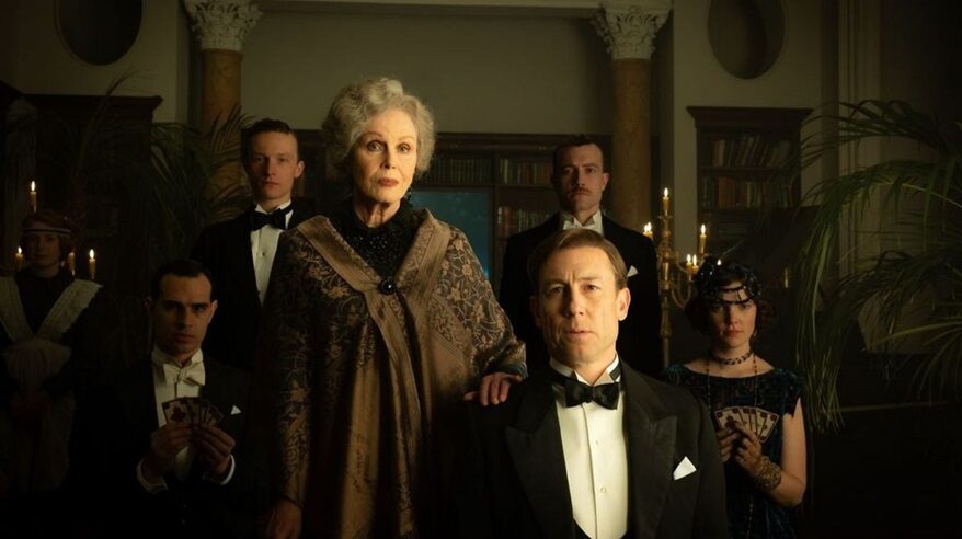 A woman stands behind a seated man, both in 19th century period costume, in a grand, dark room with others in the background