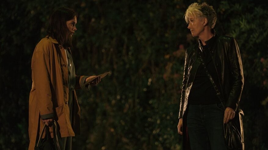 Two women stand facing each other in the dark with bushes behind them