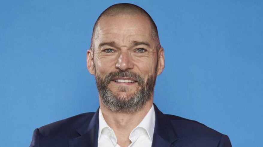 A man with close-shaved hair and beard ion white shirt and blue jacket looks to camera with pale blue background
