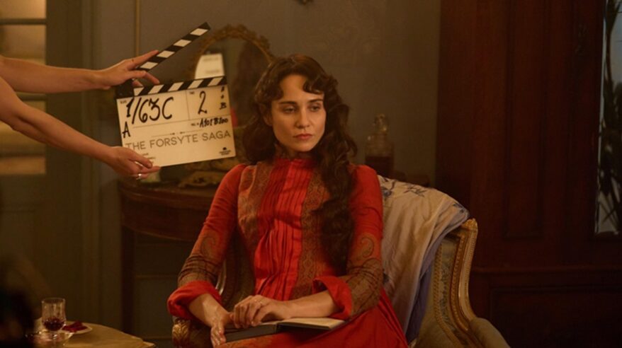 Lady in 19thC red dress sits on chair with film clapperboard to her right side