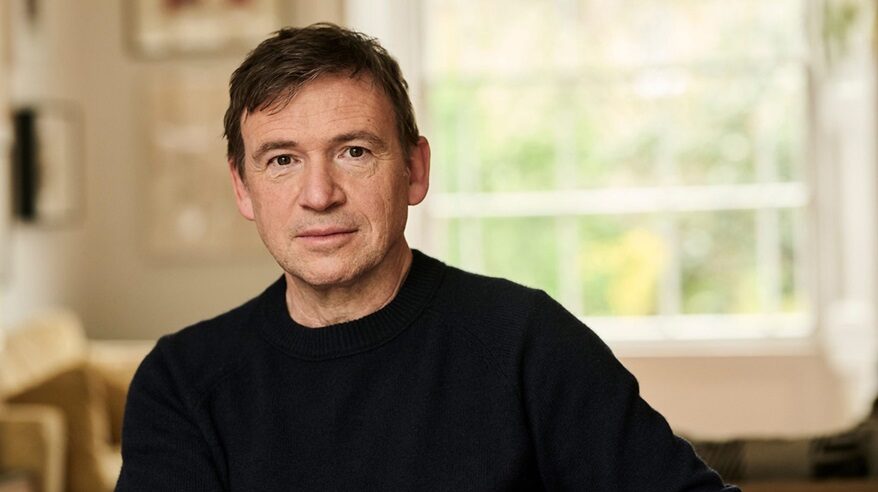 A dark-haired man in a dark sweater looks out to camera, with a window behind him